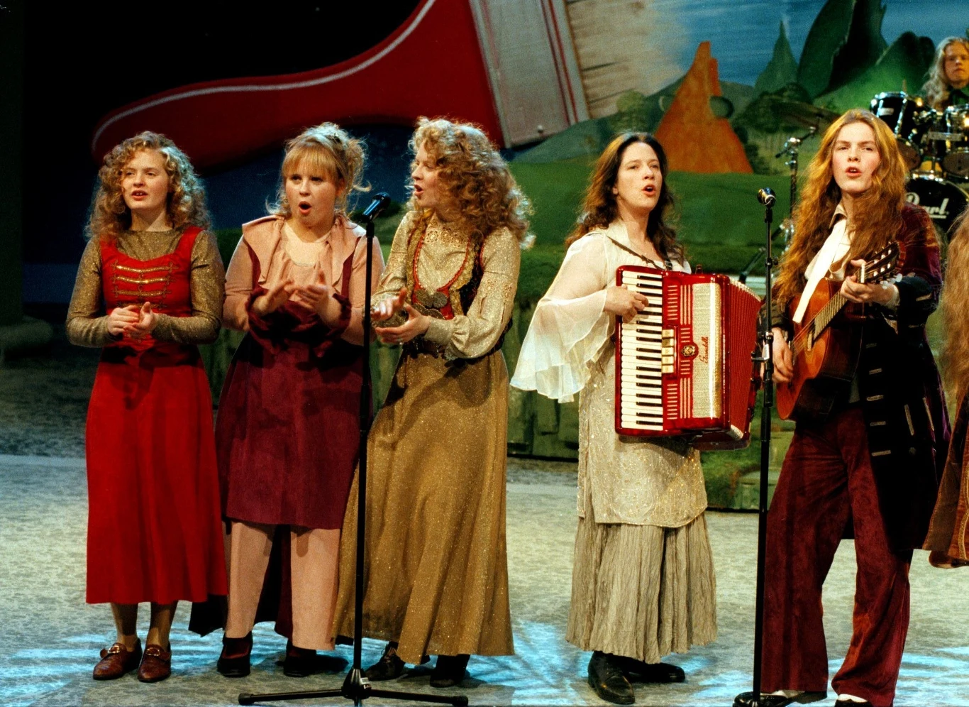 The Kelly Family: Barby, Maite, Patricia, Kathy i Paddy Kelly Grupa pięciu kobiet na scenie, wszystkie ubrane w stylowe ubrania w stylu retro, śpiewają i grają na instrumentach, w tym akordeonie i gitarze. W tle widoczna jest scenografia przypominająca pejzaż z monumentalnym fortepianem.