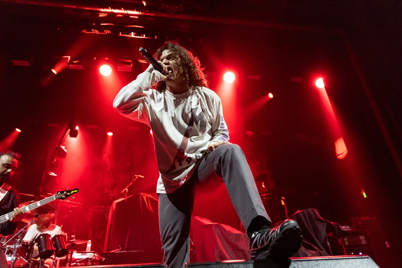 Simon Olsen stoi na czele Baest Wokalista zespołu rockowego energetycznie śpiewa na scenie, oświetlony czerwoną poświatą świateł. W tle widać muzyków z gitarą i perkusją, tworzących dynamiczną atmosferę koncertu.