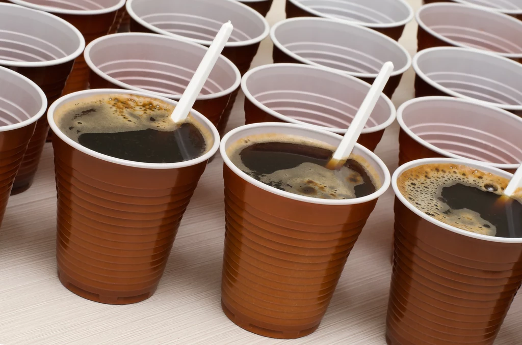 Jednorazowe opakowania plastikowe na kawę, mleko i cukier mają zostać całkowicie wycofane Plastikowe kubki wypełnione czarną kawą, każdy z plastikową łyżeczką. Kubki układają się w kilku rzędach na stole.