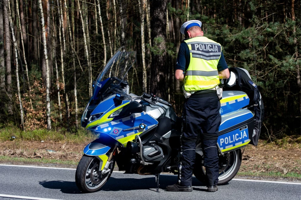 Policjant w kamizelce odblaskowej stoi obok motocykla policyjnego na drodze, otoczony lasem. Motocykl jest oznaczony napisem 'Policja'. Scena sugeruje rutynową kontrolę drogową lub patrol w spokojnym, zalesionym terenie.