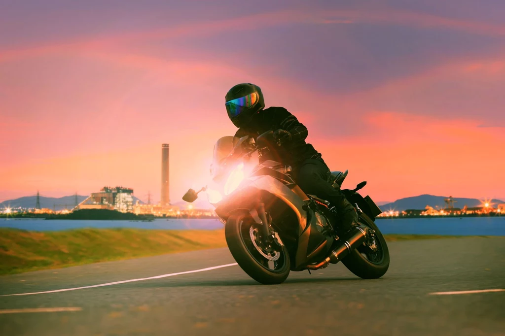 Motocyklista w stroju ochronnym i kasku jadący na motocyklu po pustej drodze o zachodzie słońca. W tle widoczna jest zabudowa przemysłowa oraz piękne, kolorowe niebo.