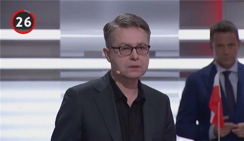 Maciej Maciak potwierdził, że podziwia Władimira Putina Mężczyzna w okularach i garniturze stoi na tle jasnego, nowoczesnego studia telewizyjnego, z mikrofonem przypiętym do ucha. Obok, w tle, inny uczestnik trzyma małą flagę.