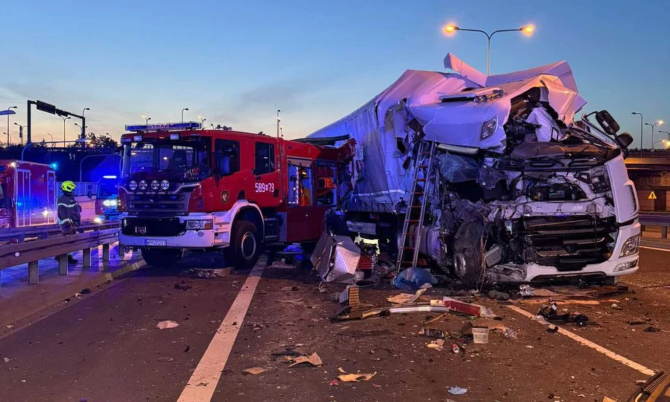 Warszawa Węzeł Lotnisko. Wypadek z udziałem wozu strażackiego Wypadek drogowy na autostradzie, z udziałem ciężarówki i wozu strażackiego. Pojazdy są mocno uszkodzone, wokół walają się szczątki. W tle widać wieczorne niebo oraz oświetlenie uliczne.