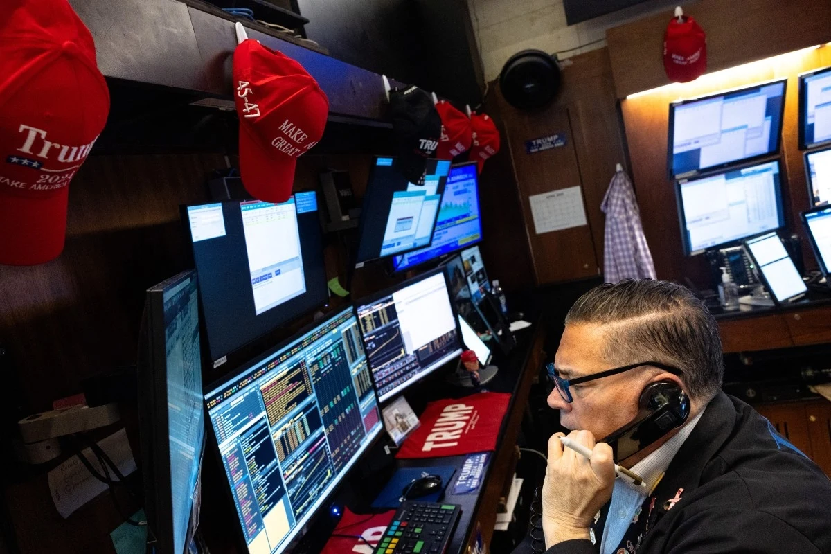 USA. Wall Street - tak wygląda praca w nowojorskiej giełdzie Osoba siedząca przy monitorach komputerowych z widocznymi czapkami z napisem 'Trump' i 'Make America Great Again', w otoczeniu ekranów pokazujących dane i wykresy.