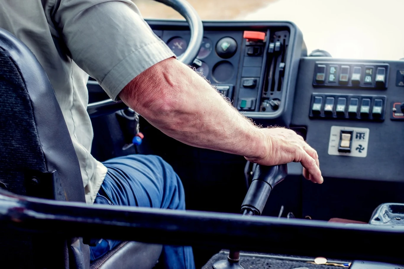 Ręka mężczyzny trzymająca dźwignię w kabinie pojazdu z widocznymi przyciskami i wskaźnikami na panelu kontrolnym w tle.