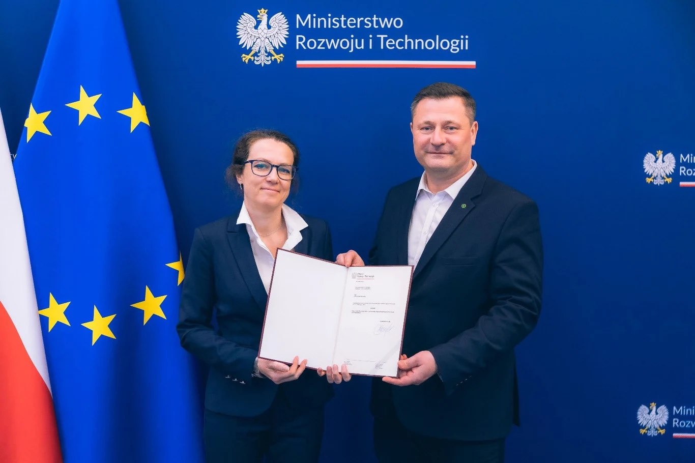 Dr Marta Ewa Wachowicz została nową prezes Polskiej Agencji Kosmicznej Mężczyzna i kobieta w formalnych strojach stoją na tle granatowej ściany z logiem Ministerstwa Rozwoju i Technologii, trzymając wspólnie dokument. Obok flagi Polski i Unii Europejskiej.