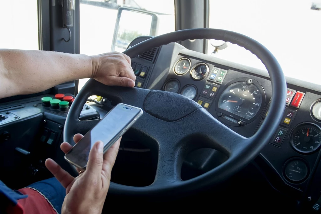 Za korzystanie z telefonu podczas jazdy grożą surowe kary. Kierowca siedzi za kierownicą dużego pojazdu, trzymając w jednej ręce telefon komórkowy. Z instrumentów na desce rozdzielczej widoczne są wskaźniki i przyciski panelu sterowania w kabinie pojazdu.