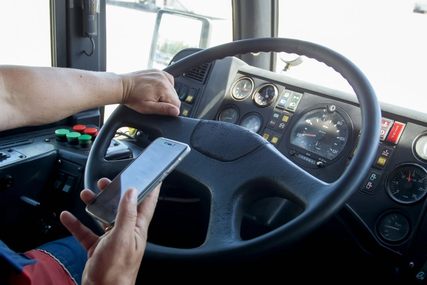 Kierowca siedzi za kierownicą dużego pojazdu, trzymając w jednej ręce telefon komórkowy. Z instrumentów na desce rozdzielczej widoczne są wskaźniki i przyciski panelu sterowania w kabinie pojazdu.