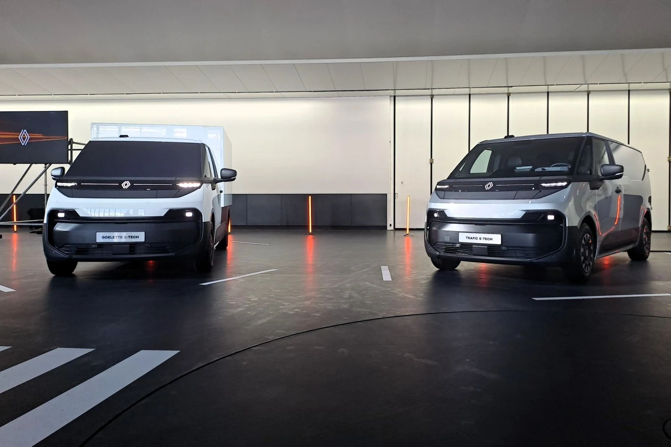 Dwa nowoczesne samochody dostawcze stojące obok siebie w dobrze oświetlonym, futurystycznym wnętrzu. Oba pojazdy są w ciemnym kolorze z jasnymi reflektorami. W tle widoczna jest minimalistyczna dekoracja z pionowymi liniami.