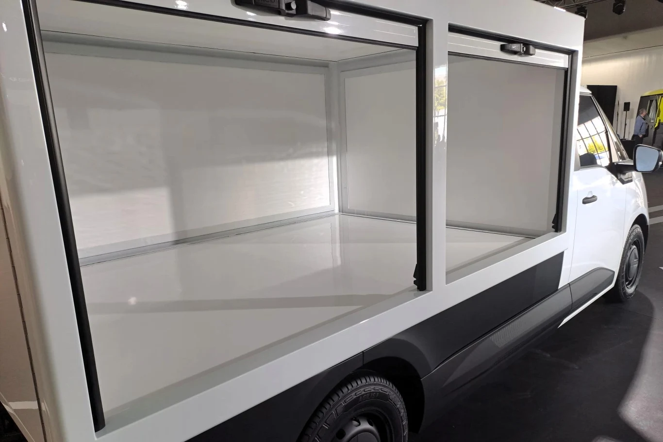 Przestronna, pusta przestrzeń transportowa wewnątrz pojazdu z dużymi oknami, pozwala na prezentację zawartości. Ściany są jasne i czyste, co sugeruje możliwość użycia do transportu lub ekspozycji produktów.
