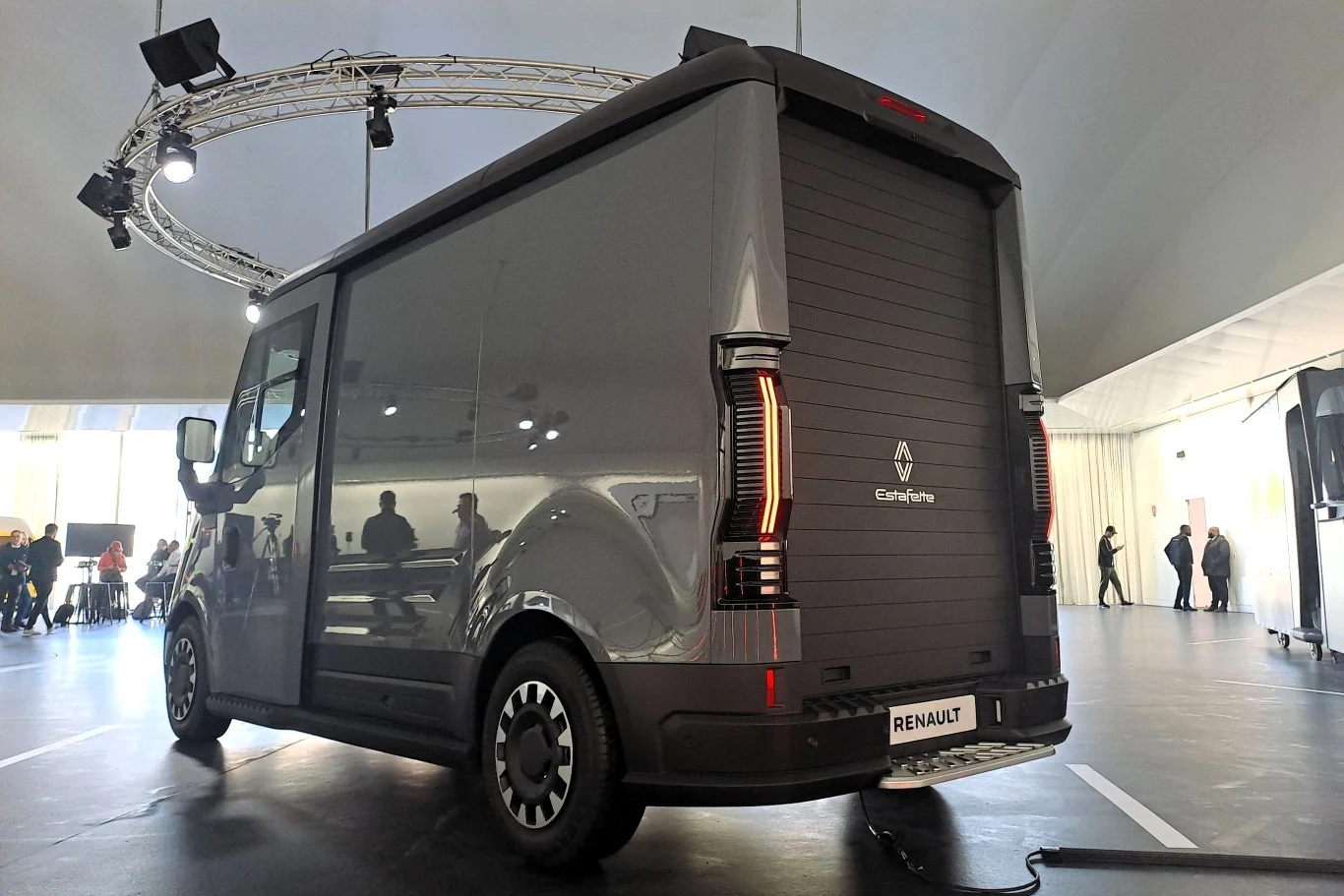 Nowoczesny van Renault o opływowych kształtach i ciemnoszarej powierzchni, stojący w pomieszczeniu oświetlonym zaawansowanym systemem świetlnym. W tle kilka osób ogląda pojazd.