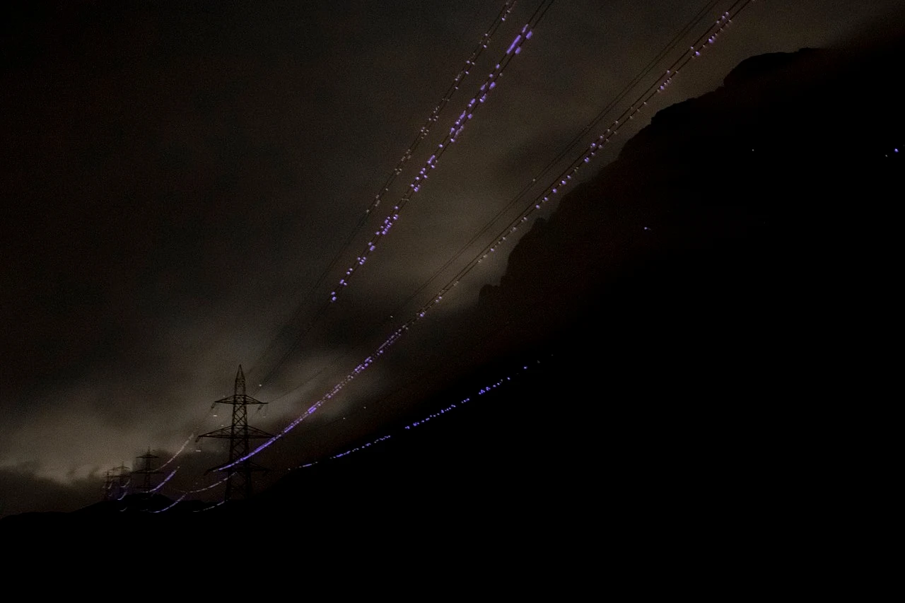 Ciemne nocne niebo z widocznymi liniami energetycznymi oświetlonymi delikatnymi świetlnymi smugami, które tworzą efektowne wzory. Po lewej stronie widać kontur pojedynczego słupa energetycznego, a w tle majaczy zarys górskiego krajobrazu.