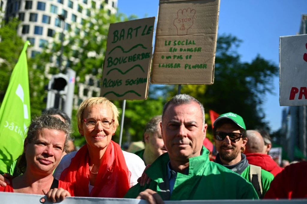 Grupa osób uczestnicząca w demonstracji trzymająca transparenty. Uczestnicy są ubrani w czerwone i zielone stroje, niektórzy mają na sobie kamizelki z nazwami organizacji. W tle widoczne są budynki i drzewa.