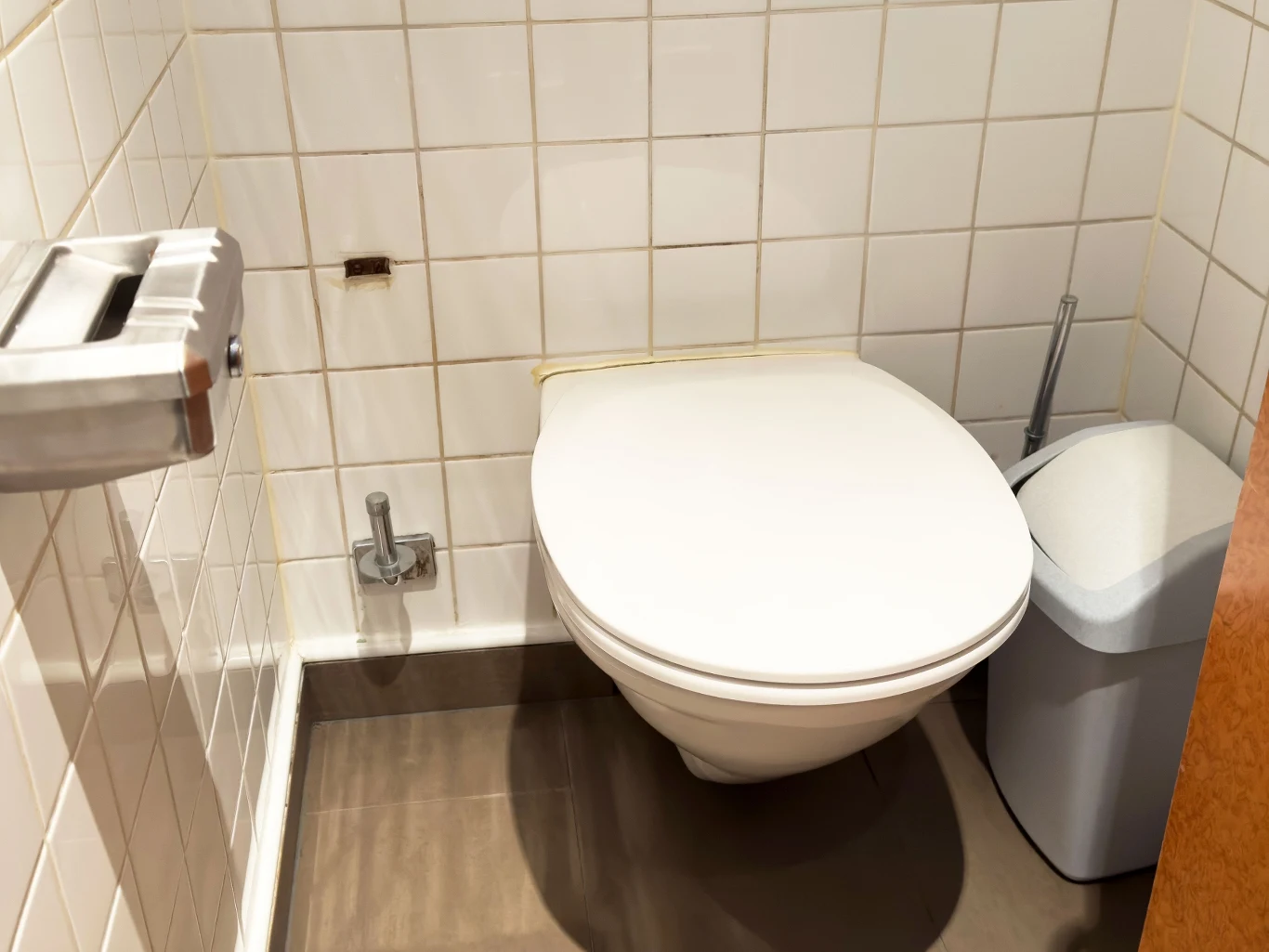 Nowoczesna i schludna łazienka z muszlą klozetową na tle jasno wyłożonej kafelkami ściany, obok której znajduje się mały kosz na śmieci oraz uchwyt na papier toaletowy.