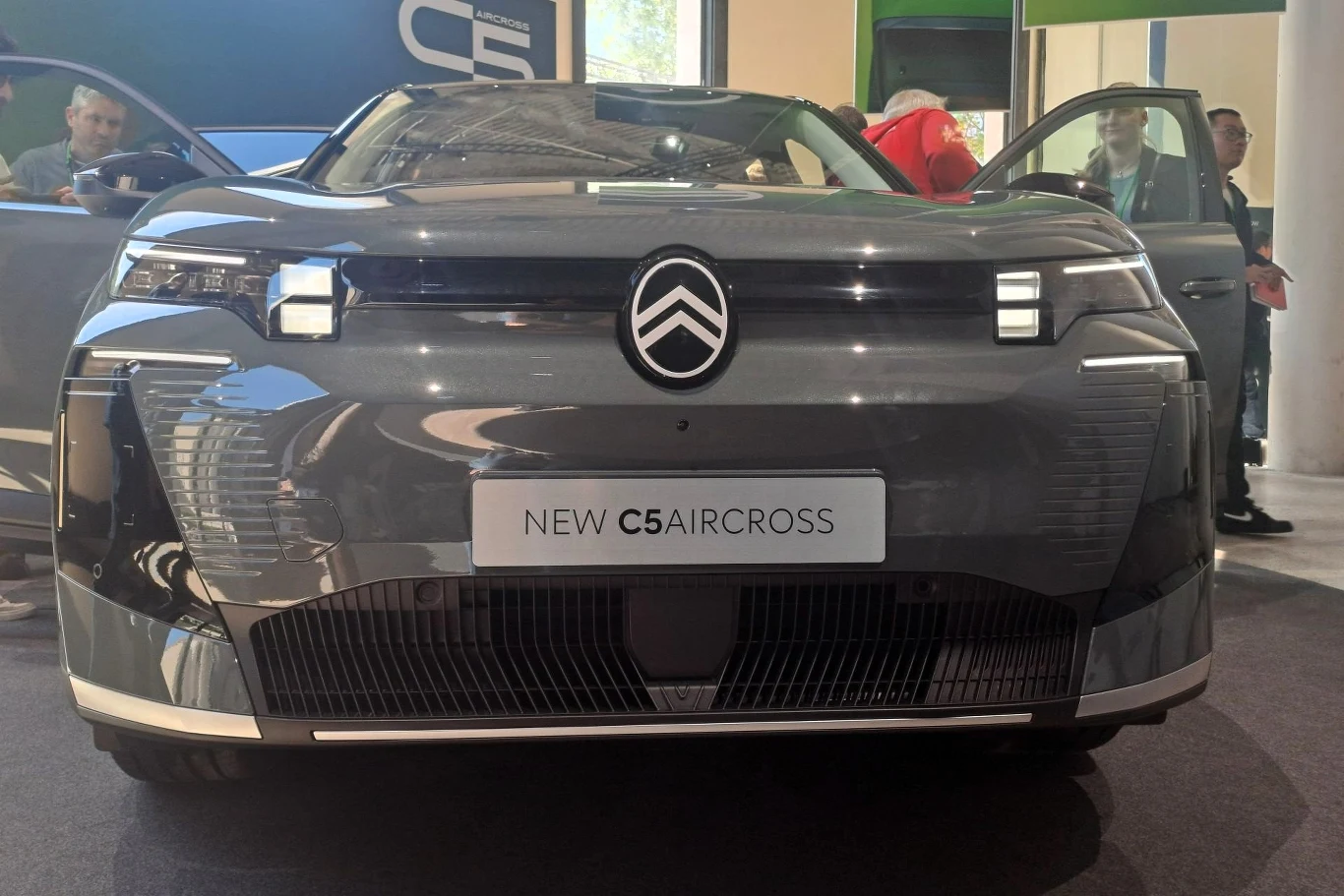 Przód szarego samochodu osobowego Citroen C5 Aircross stoi na podłodze wystawowej. Logo marki widoczne na masce, a reflektory LED podkreślają nowoczesny design pojazdu. W tle widoczni są ludzie oraz otwarte drzwi samochodu.