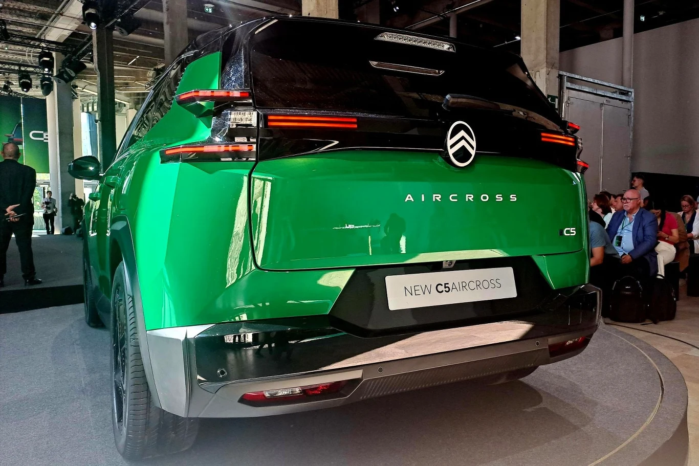 Tył zielonego samochodu Citroën C5 Aircross na wystawie motoryzacyjnej, w dobrze oświetlonym pomieszczeniu. W tle ludzie siedzący na krzesłach, rozmawiający lub obserwujący wydarzenie.