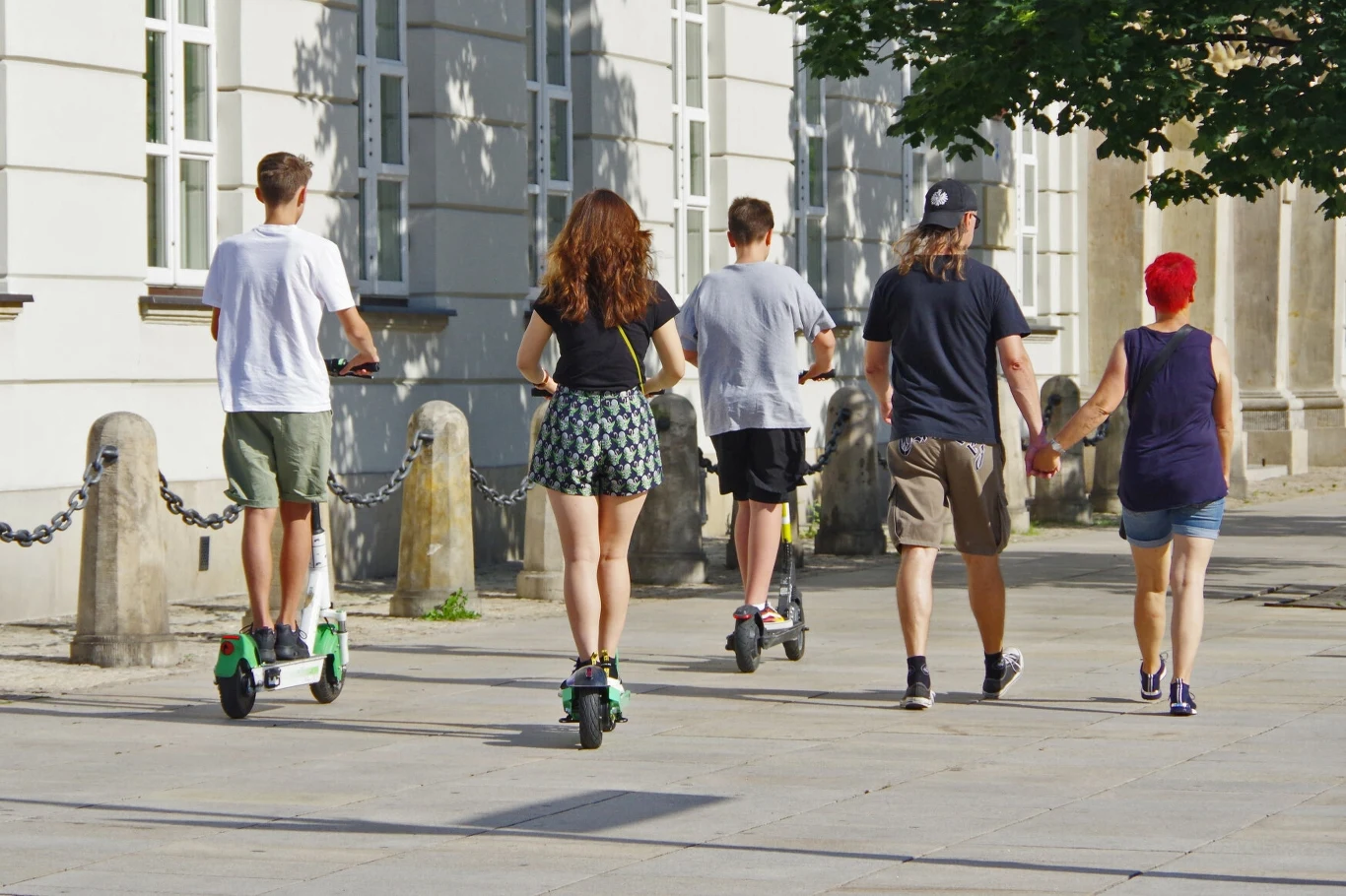 Zasady poruszania się elektrycznymi hulajnogami po chodnikach oraz wyjątki, kiedy jest to w ogóle dozwolone, regulują już obecne przepisy Grupa młodych ludzi na spacerze, dwie osoby jadą na elektrycznych hulajnogach, inne idą pieszo, tło stanowi fasada miejskiego budynku z oknami.