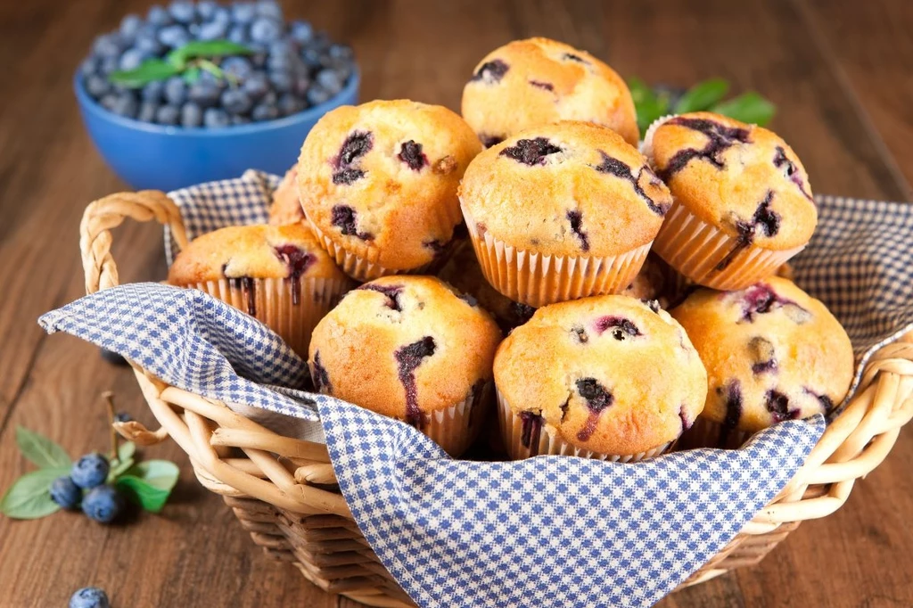 Koszyk pełen muffinek z jagodami w otoczeniu niebieskiej serwetki na drewnianym stole. W tle widoczna miseczka pełna świeżych jagód.