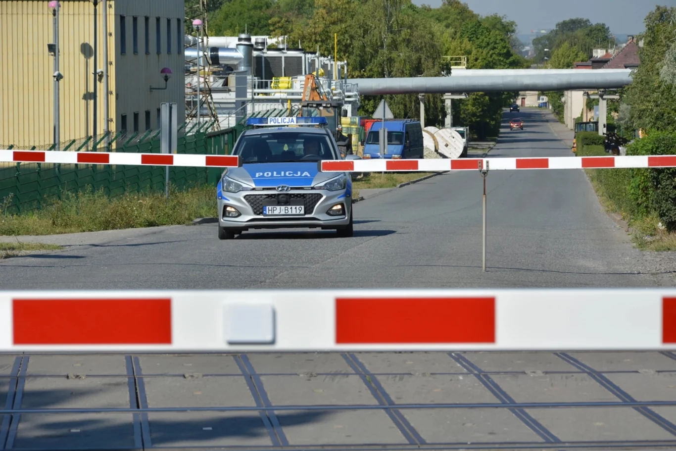 Kierowca nie może wjeżdżać na przejazd kolejowy nie tylko przy zamkniętych rogatkach, ale także w sytuacji, gdy nie są one całkowicie podniesione Samochód policyjny stoi zaparkowany za szlabanem na ulicy przemysłowej, otoczonej zielenią. Widoczne są elementy infrastruktury przemysłowej i linię torowiska na pierwszym planie.