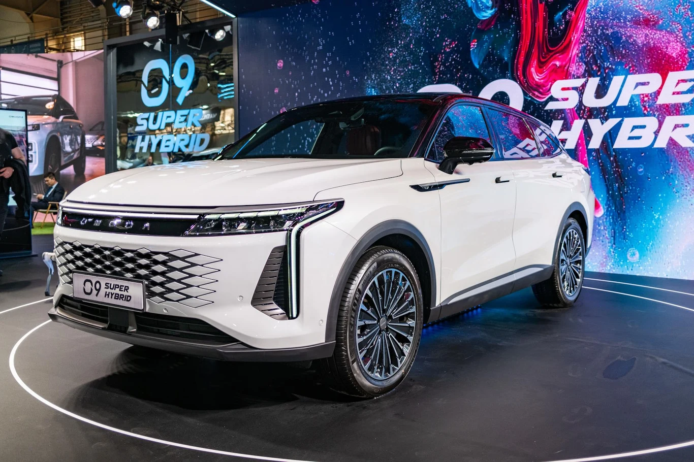 Podczas tegorocznych targów Poznań Motor Show zadebiutował flagowy SUV - OMODA 9 Super Hybrid /fot. INTERIA Biały SUV hybrydowy o nowoczesnym designie na wystawie motoryzacyjnej. Auto stoi na platformie, a w tle znajduje się duży ekran z grafiką promującą model oraz napis 'Super Hybrid'. Oświetlenie podkreśla eleganckie linie pojazdu.