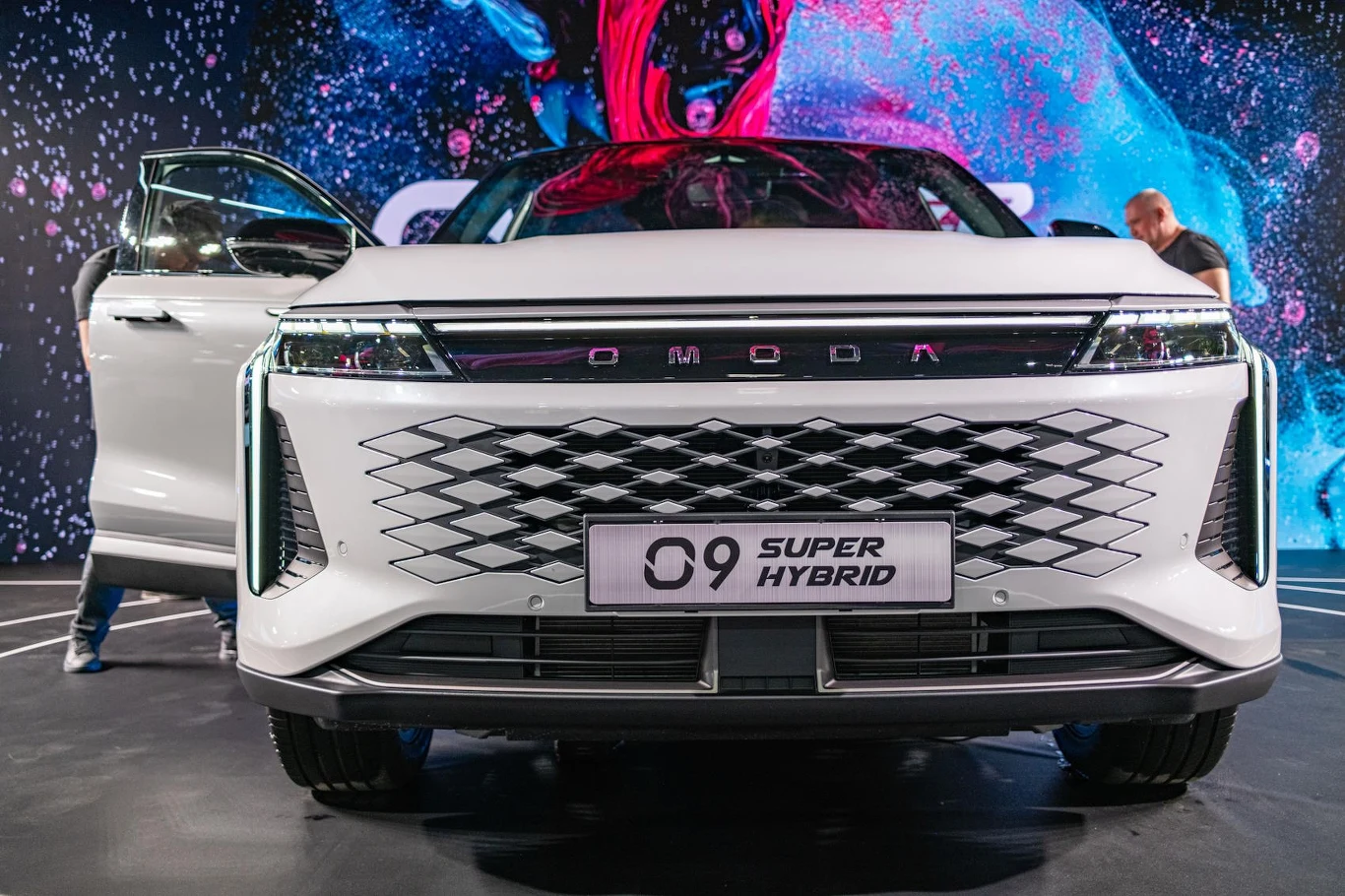 Nadwozie wyróżnia się nowoczesnymi proporcjami /fot. INTERIA Biały samochód OMODA O9 Super Hybrid z otwartymi drzwiami, widoczny od przodu, stoi na tle kolorowej ściany LED. Przednia krata z charakterystycznym wzorem i tablica rejestracyjna z nazwą modelu dominują w kompozycji.