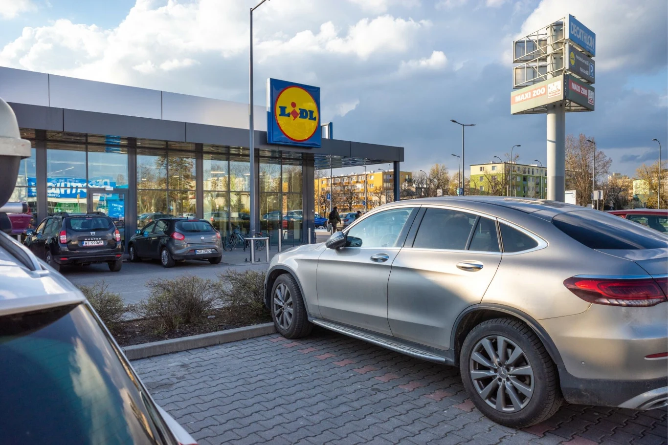 Parking Lidl - kamery zamiast parkometrów. Jak działa nowy system? Wejście do supermarketu Lidl z zaparkowanymi samochodami przed budynkiem. Na pierwszym planie samochód osobowy, w tle budynek sklepu i niebo z chmurami.