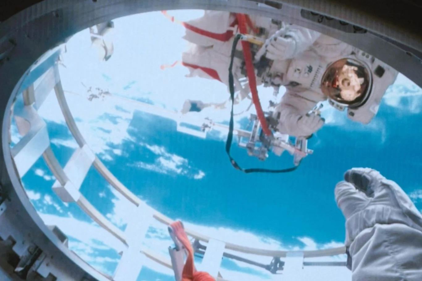 Chińscy tajkonauci Cai Xuzhe (u góry) i Song Lingdong podczas spaceru kosmicznego Astronauta w skafandrze kosmicznym wykonuje czynności poza stacją kosmiczną, widoczny jest niebieski horyzont Ziemi. Przez okrągły otwór widać dłoń w rękawicy trzymającą narzędzie.”