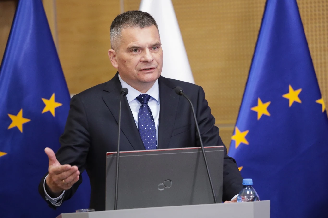 Prokuratura Krajowa przedstawiła drugą część raportu z audytu Mężczyzna w garniturze i krawacie przemawiający przy mównicy, z dwoma mikrofonami przed sobą. W tle flagi Unii Europejskiej oraz flaga biała.