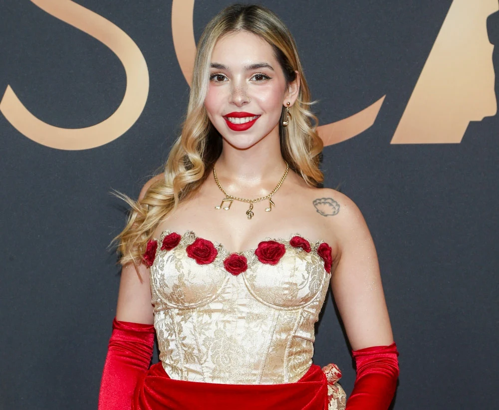 Aria Martelle prezentuje nową piosenkę Kobieta w eleganckiej sukni z gorsetem ozdobionym czerwonymi różami. Suknia w kolorze złotym i czerwonym. Uśmiechnięta, z długimi falującymi włosami, stoi na tle czarno-złotego napisu.