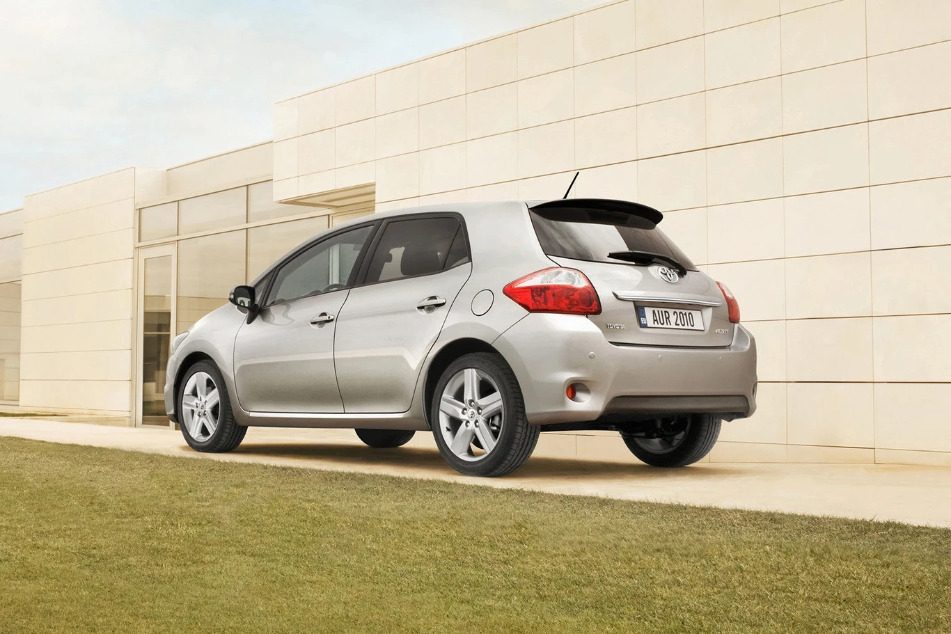Toyota Auris to wciąż hit na rynku wtórnym, Srebrny samochód osobowy zaparkowany pod nowoczesnym, jasnym budynkiem o prostych liniach architektonicznych. Pojazd znajduje się na trawniku, a w tle widać ściany ze szkła i jasnego kamienia.