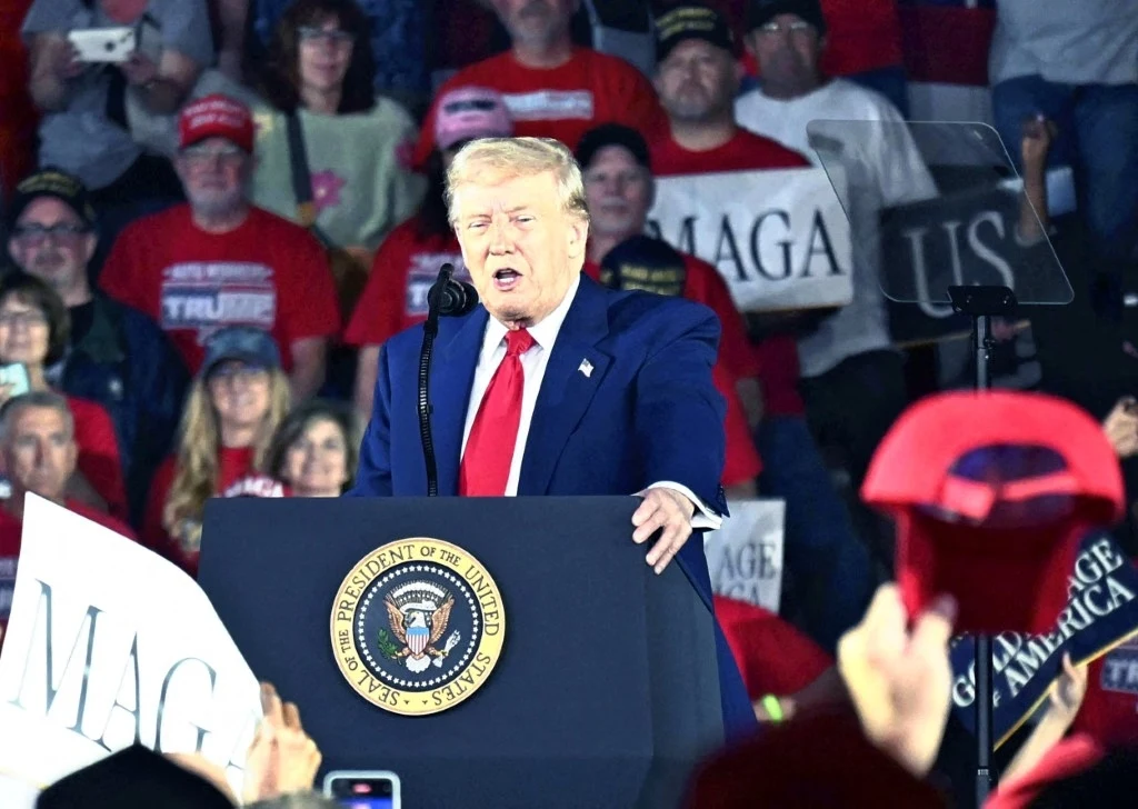 Donald Trump podczas przemówienia z okazji 100 dni prezydentury Były prezydent USA wygłasza przemówienie na wiecu, ubrany w niebieski garnitur i czerwoną krawat. W tle tłum ludzi trzymających transparenty z napisem MAGA.