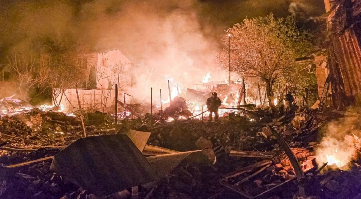 Skutki ataku na obwód charkowski Scena pełna gruzów i płomieni, z mężczyznami w tle próbującymi poruszać się przez chaos zniszczeń. Drzewa i fragmenty budynków wśród unoszącego się dymu wskazują na dużą skalę zniszczeń.
