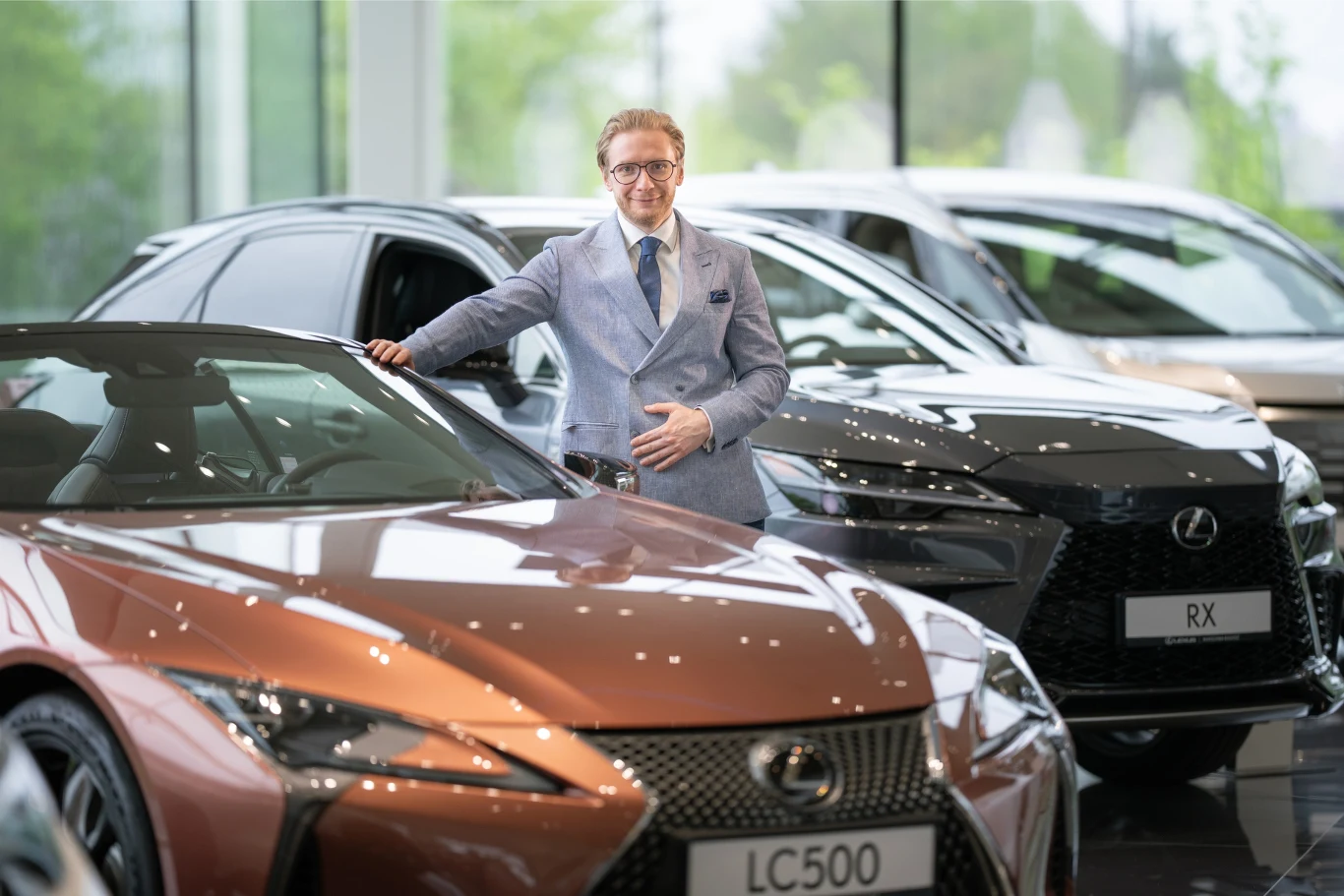 Mężczyzna w eleganckim garniturze stoi obok luksusowego samochodu marki Lexus w salonie samochodowym. Przedstawione są modele LC500 i RX, które wyróżniają się nowoczesnym wzornictwem. W tle widoczne są inne samochody.