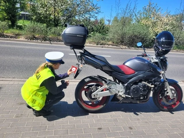 Policjantka w żółtej kamizelce odblaskowej kontroluje motocykl zaparkowany na chodniku. Obok niej położony jest identyfikator pojazdu oparty o tylne koło motocykla. W tle widoczna jest droga oraz drzewa.