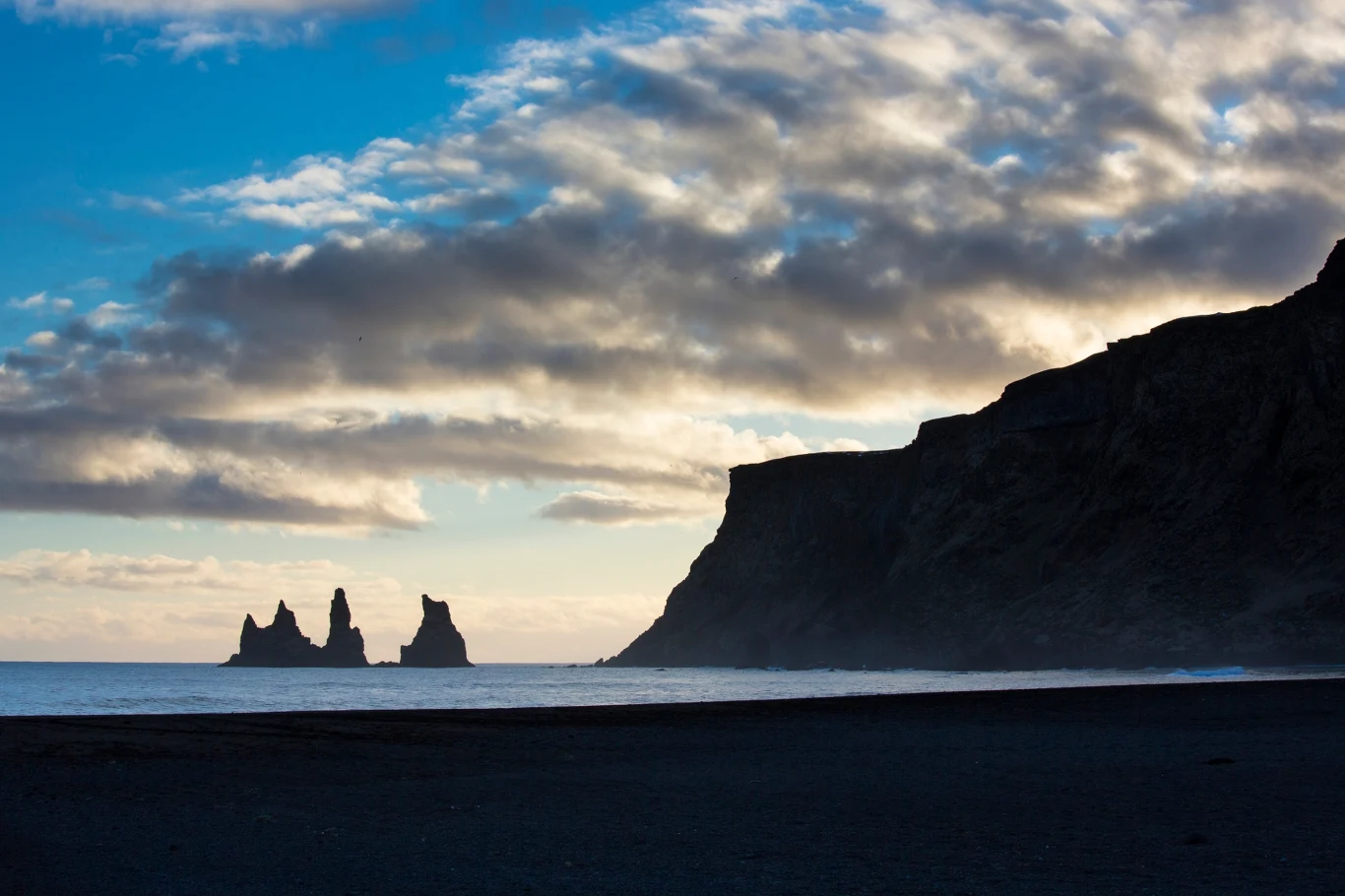 Plaża Reynisfjara na Islandii Widok na ciemną plażę z widocznymi w oddali charakterystycznymi formacjami skalnymi w oceanie, przy zachmurzonym niebie, które tworzy dramatyczny klimat.