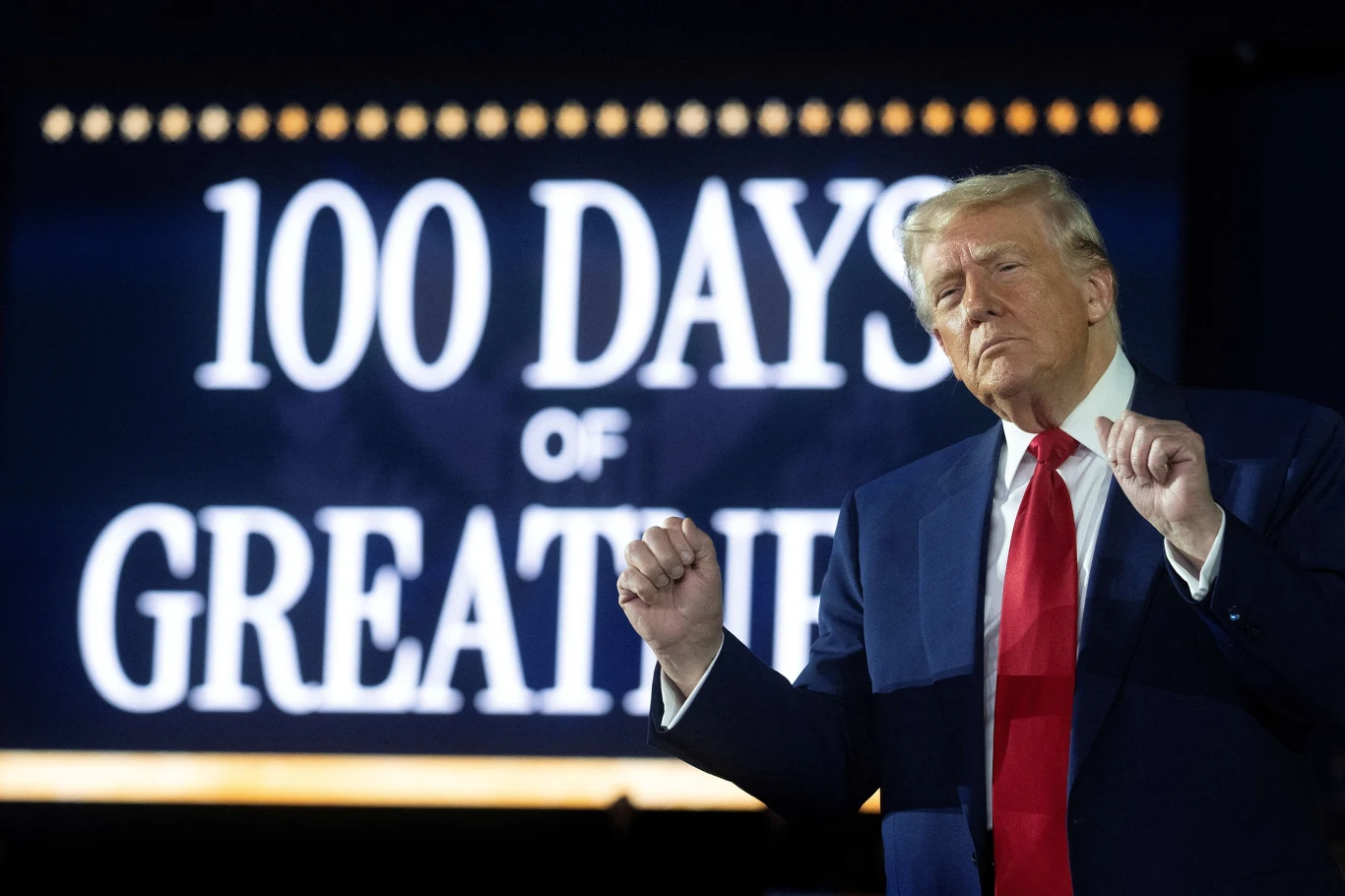 Donald Trump nie traci dobrego samopoczucia Mężczyzna w garniturze z czerwoną krawatą stoi na tle dużego napisu '100 DAYS OF GREATNESS'. Jego wyraz twarzy i gesty sugerują jakość publicznego wystąpienia lub prezentacji.