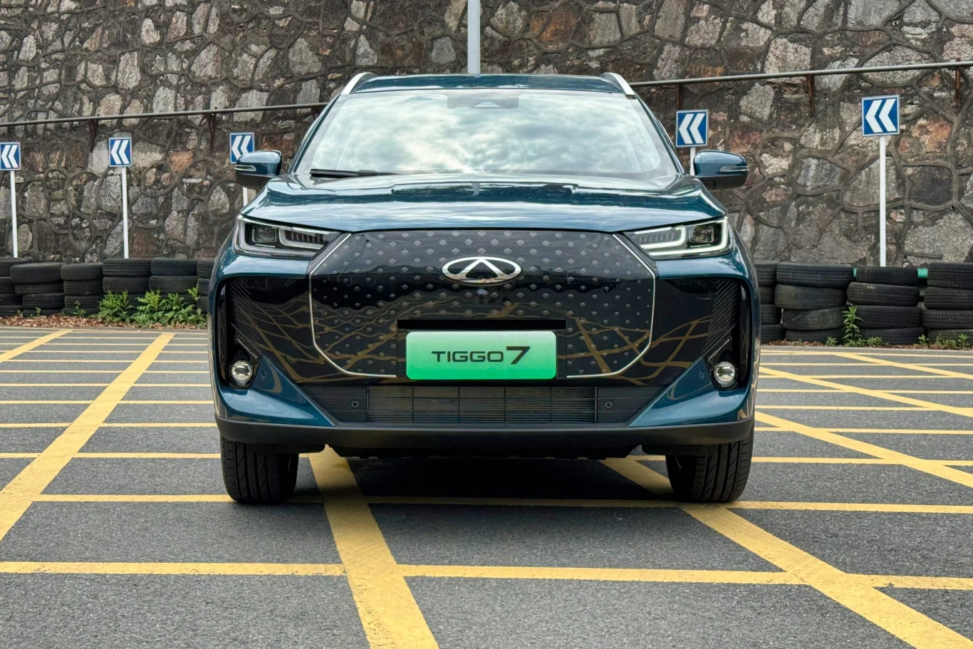 Samochód Chery Tiggo 7 o nowoczesnym designie stoi zaparkowany na parkingu z żółtymi liniami. Przód auta jest dobrze widoczny, z wyraźnym logo marki i stylowym grillem. Tło tworzy mur z kamienia, a w rogu widać stos opon.