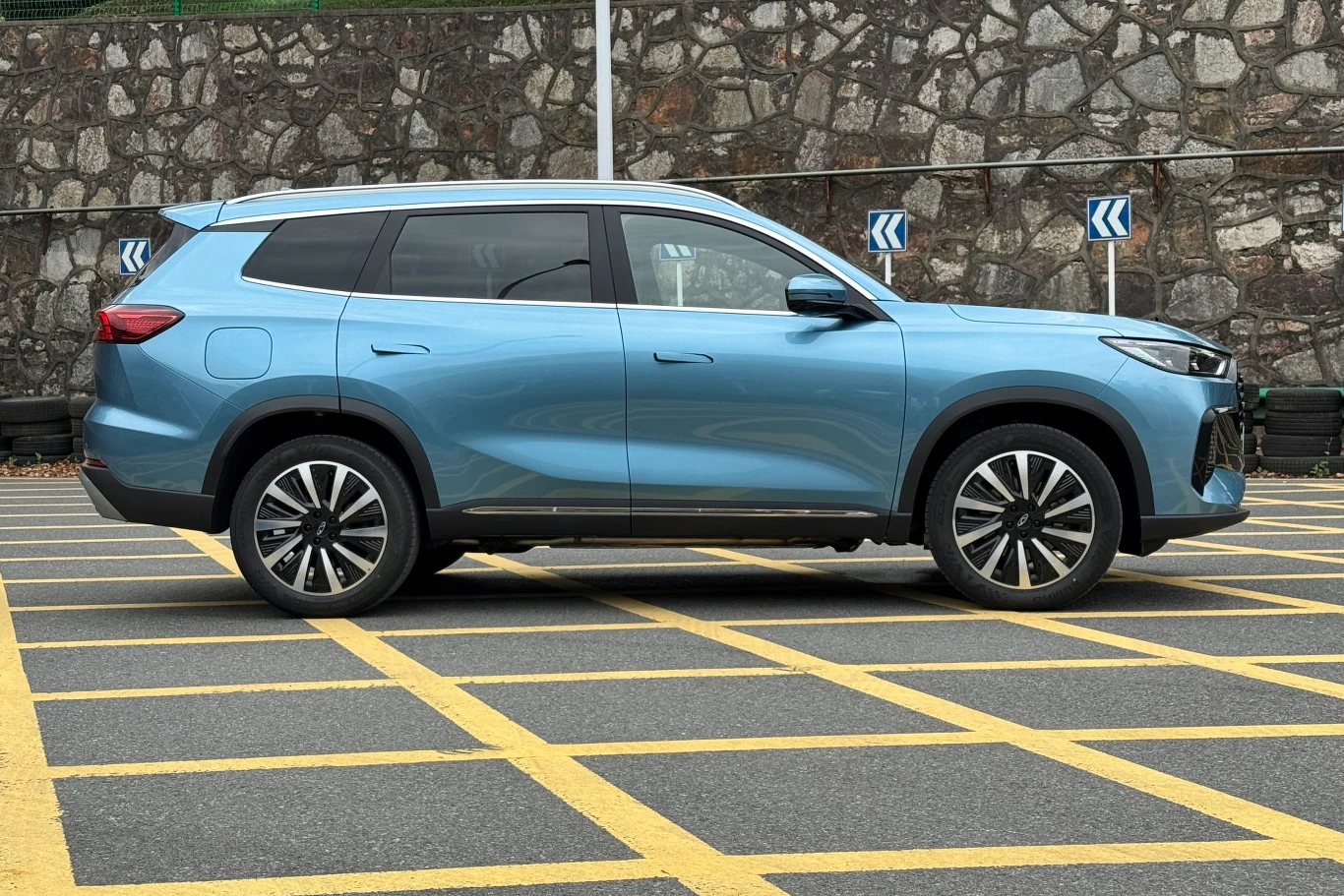 Chery Tiggo 8 zadebiutuje w Polsce już 30 czerwca Niebieski SUV zaparkowany na parkingu z wyznaczonymi żółtymi liniami, obok kamiennego muru.