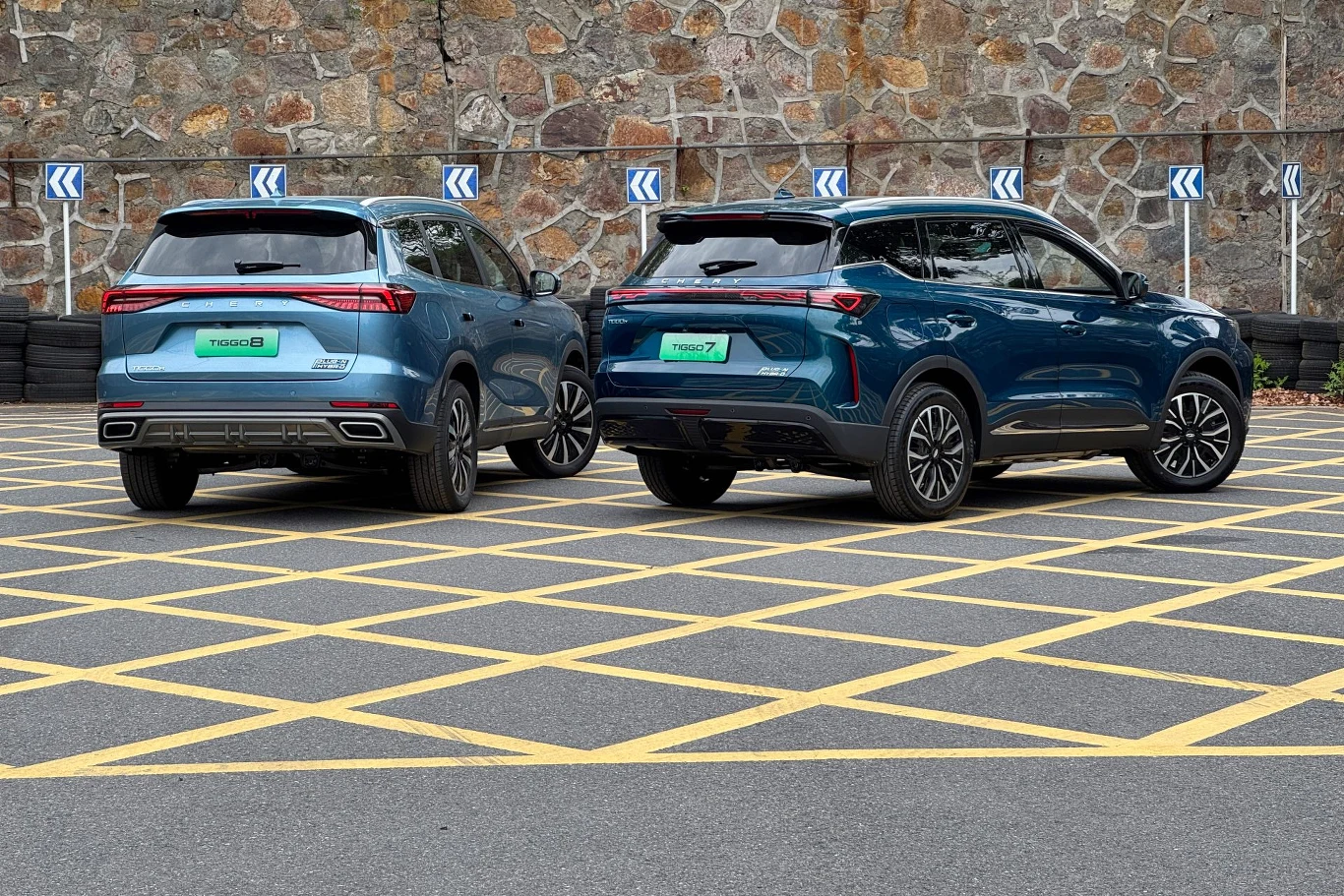 Dwa niebieskie pojazdy SUV stoją zaparkowane na pustym parkingu z żółtymi oznaczeniami linii, w tle widoczny mur z kamienia.