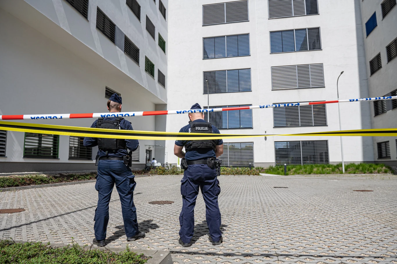 Atak na lekarza w Krakowie. 35-latek usłyszał zarzuty Dwóch policjantów stoi tyłem do obiektywu, za taśmą policyjną, przed nowoczesnym budynkiem mieszkalnym, stłumione światło dnia uwydatnia elementy architektury.