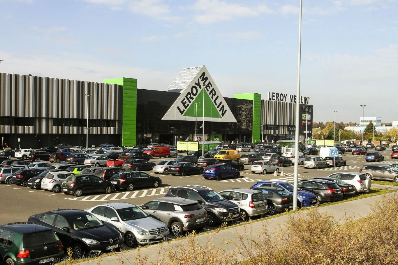 Duży parking przed budynkiem sklepu Leroy Merlin wypełniony samochodami. Budynek o nowoczesnym designie z wyraźnym logo na froncie i charakterystyczną zielono-czarną kolorystyką.