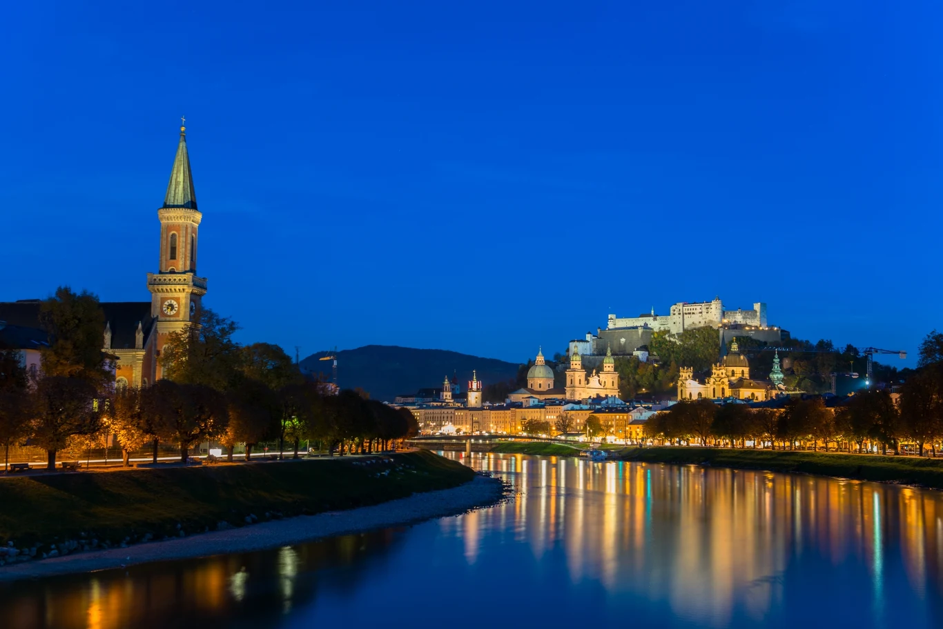Nocna panorama Salzburga z widokiem na rzekę Salzach odbijającą światła miasta. Po lewej stronie widoczny jest wysoki kościół, a w tle oświetlony zamek Hohensalzburg na wzgórzu.