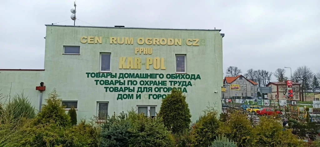 Centrum ogrodnicze w Bartoszycach. Na fasadzie widać napis w cyrylicy Budynek centrum ogrodniczego z dużym napisem w języku polskim i rosyjskim. Otoczony roślinnością, w tle widać inne budynki oraz pochmurne niebo.