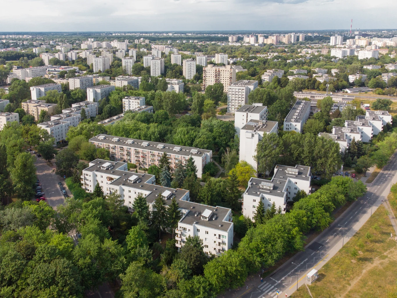 Osiedle z lat 60. XX wieku w Warszawie – przykład późnomodernistycznej architektury. Widok z lotu ptaka na miejskie blokowiska otoczone dużą ilością zieleni, w oddali widać panoramę miasta.