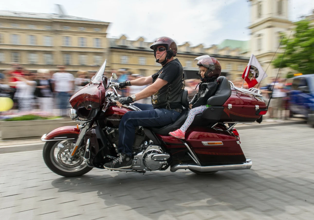 Podczas jazdy motocyklem z dzieckiem najważniejsze jest bezpieczeństwo Motocyklista wraz z dzieckiem, obaj w kaskach, jadą czerwonym motocyklem ulicą miasta. W tle zarys tłumu zebranych ludzi oraz budynki miejskie.