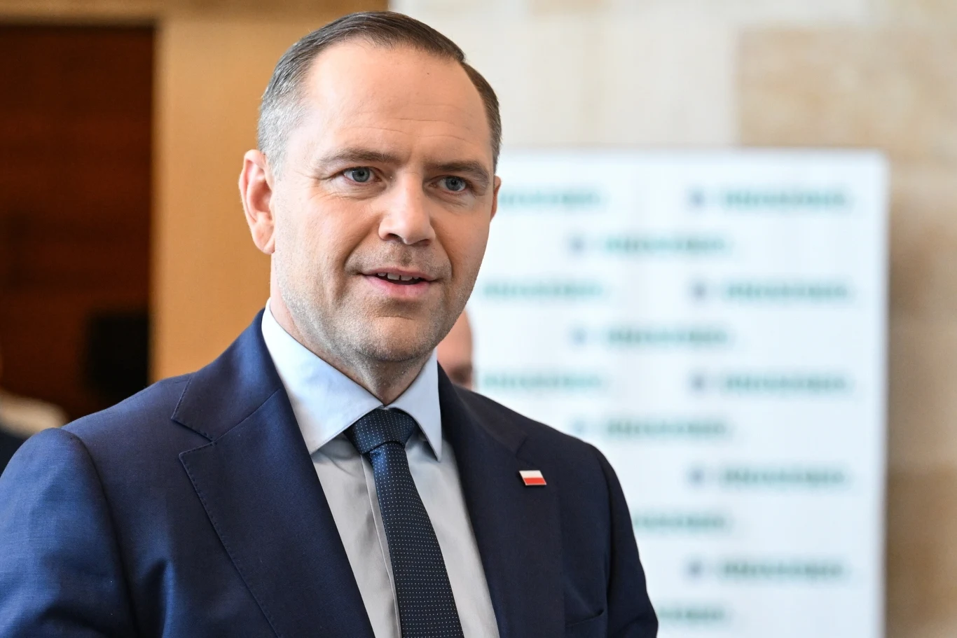 Politycy opozycji komentują medialne doniesienia ws. liczby mieszkań Karola Nawrockiego Mężczyzna w garniturze z flagą Polski na klapie stoi w formalnym otoczeniu, w tle widoczne rozmyte logo.