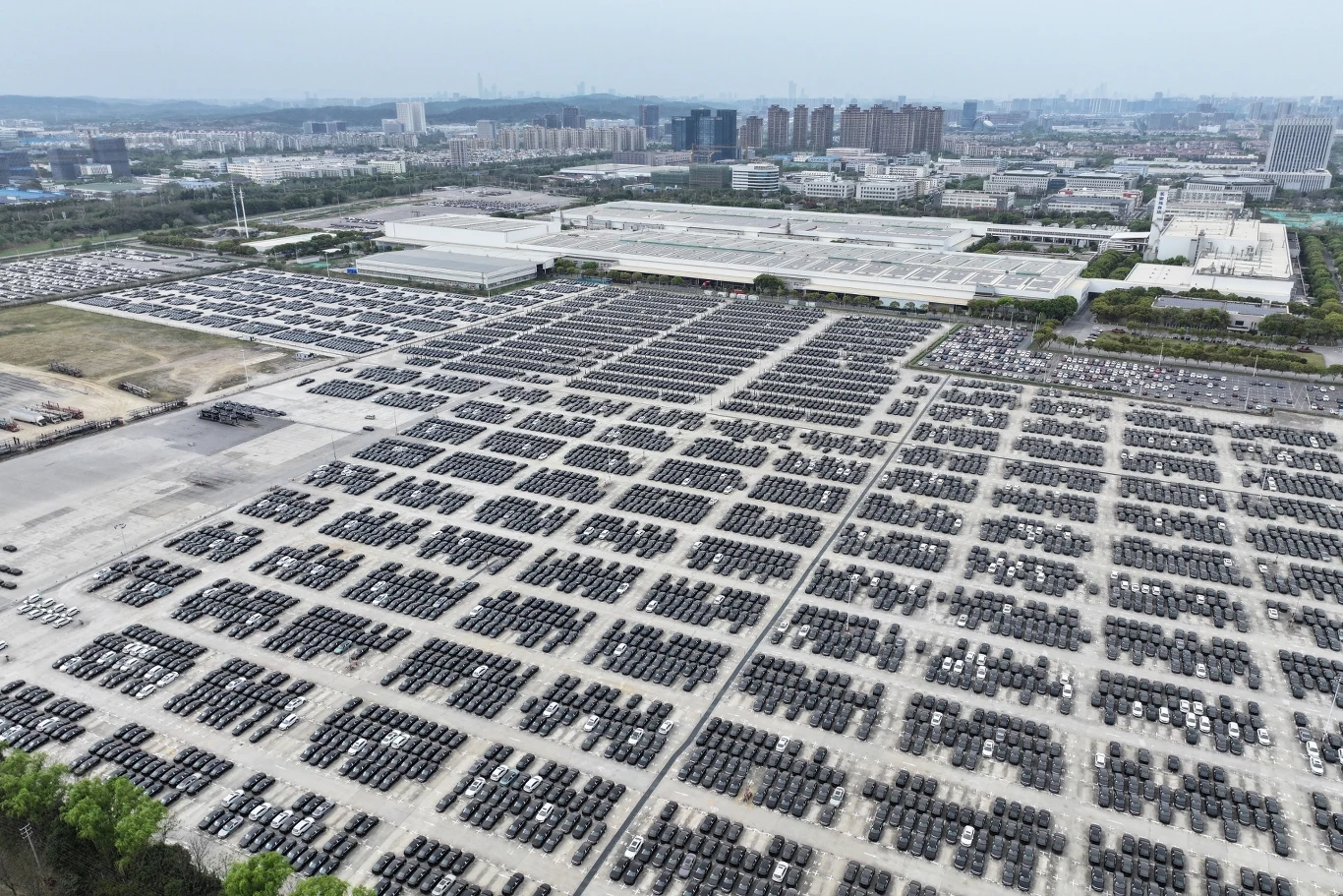Ogromny parking pełen czarnych opon samochodowych ułożonych w uporządkowanych rzędach, z fabryką i miastem w tle. Widok z lotu ptaka ukazuje skalę produkcji przemysłowej w okolicy.