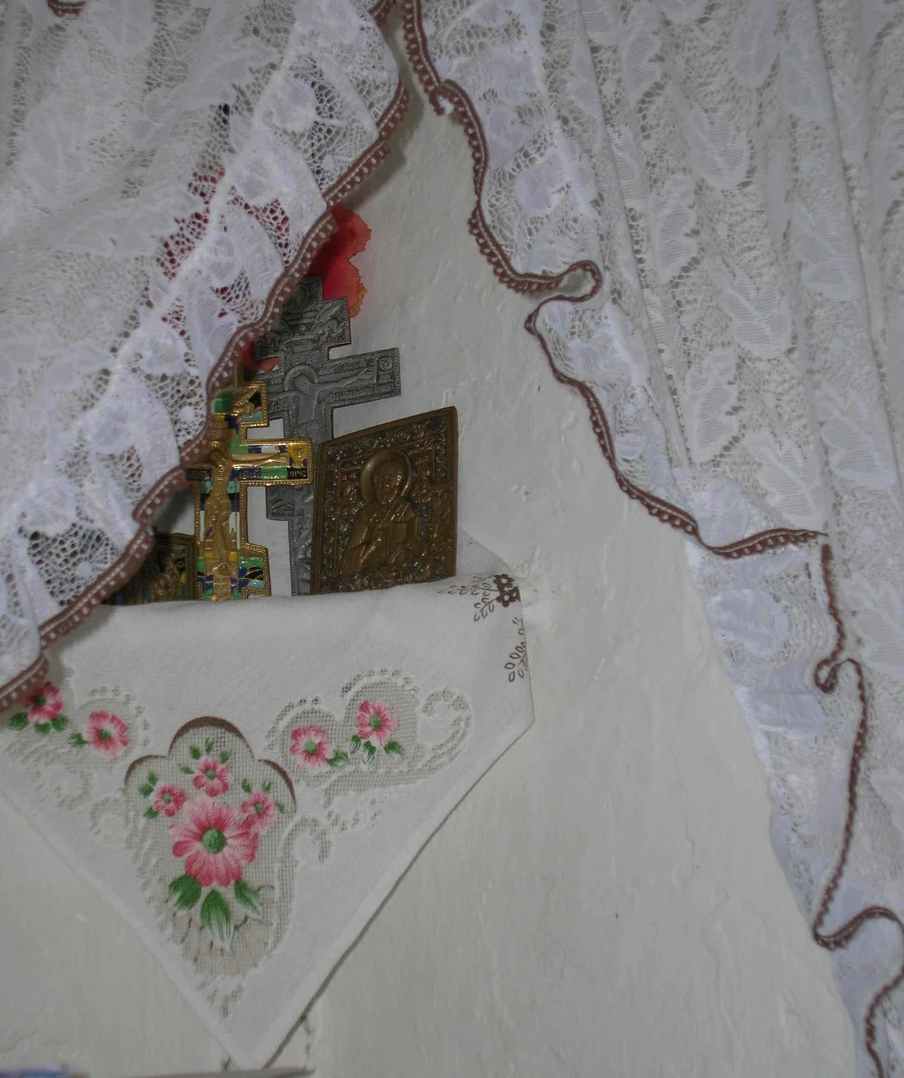 Krasny kąt z ikonami/ Fot. Katarzyna Roman-Rawska Obrazek pokazuje kącik z ikoną religijną osłoniętą dekoracyjną serwetką z kwiatowym wzorem, otoczoną koronkową firanką. Ikona i krzyż są centralnym elementem wystroju. Całość nadaje miejscu intymny, sakralny charakter.