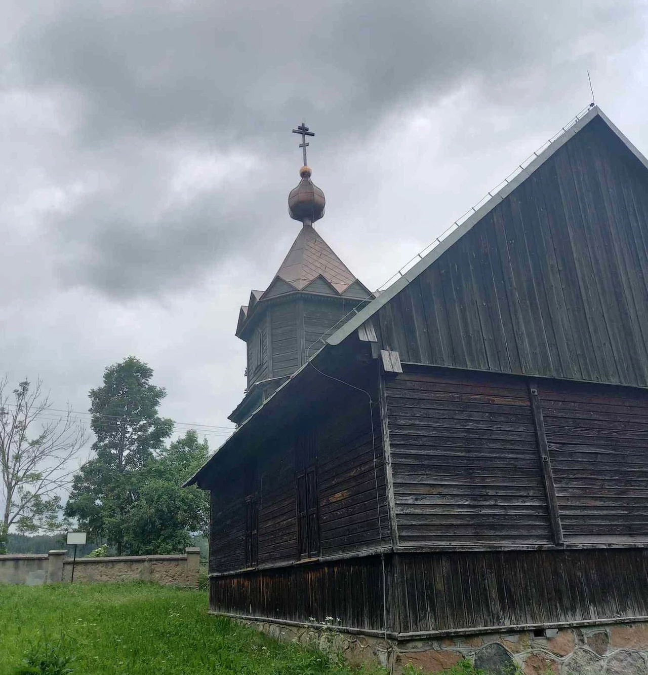 Molenna, czyli świątynia starowierców/ Fot. Katarzyna Romasn-Rawska Drewniany kościół z cebulastą kopułą, otoczony zielenią i chmurami na niebie. Konstrukcja wykonana z ciemnego drewna, osadzona na kamiennym fundamencie.