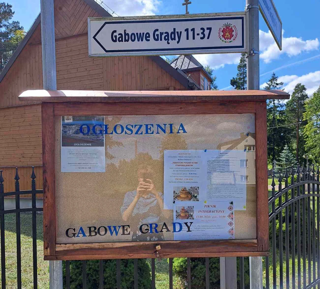 Tablica ogłoszeń w drewnianej ramie z różnymi wydrukowanymi informacjami, w tle fragment drewnianego budynku oraz znak drogowy z nazwą miejscowości Gabowe Grądy.